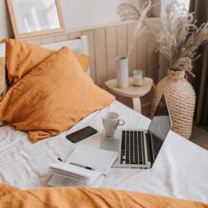 From above of open netbook with black screen and cup of coffee with smartphone and notebook placed on crumpled bed sheet in bedroom with big vase on floor