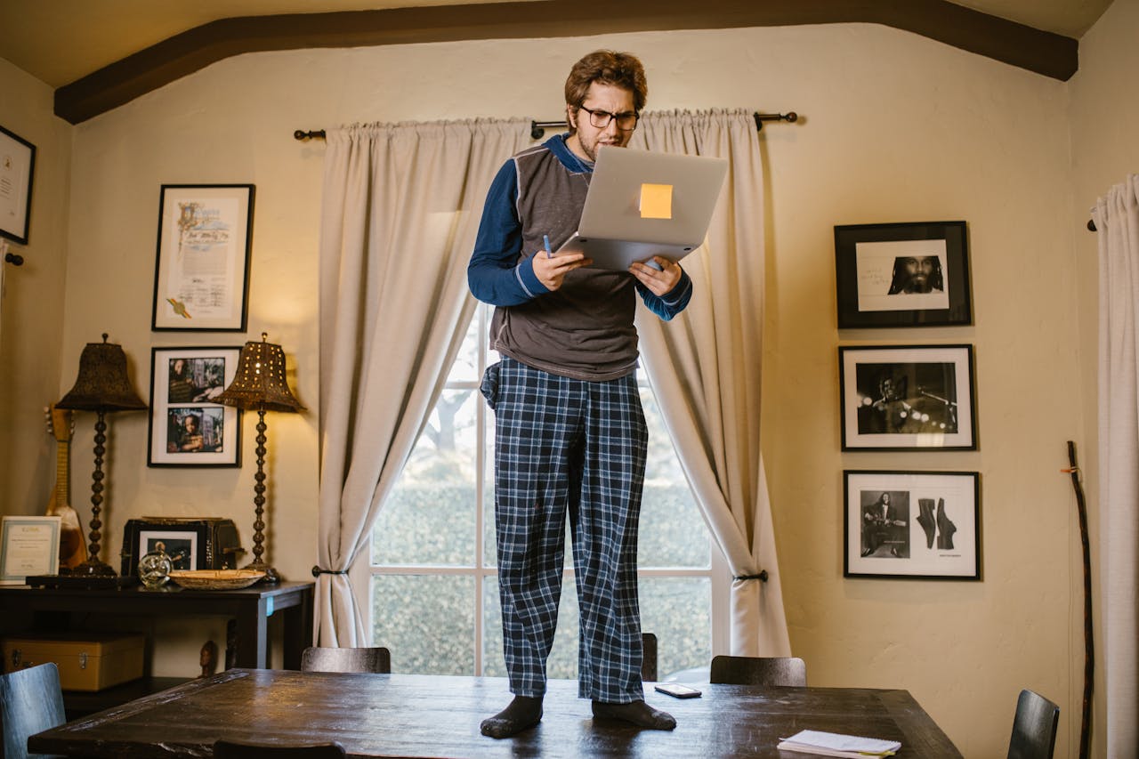 process-img Man in pajamas standing on table with a laptop indoors, focused on remote work or study.